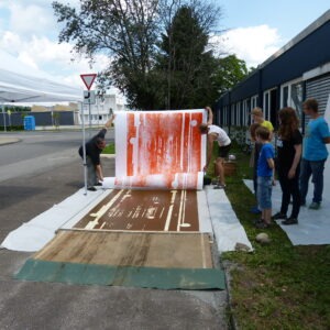 Zwei Personen lösen einen 1,50 x 2,70 Meter großen Papierbogen von der Druckplatte. Das Bildmotiv sind weiße Streifen und Kreise auf orangefarbenem Hintergrund. Am Rand stehen Kinder und Jugendliche und schauen zu